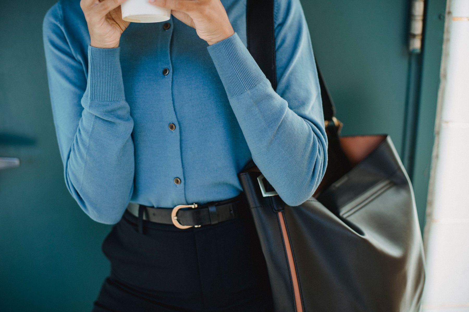 Cropped view of a person wearing a blue cardigan and black trousers with a black belt, holding a white cup and carrying a large black tote.