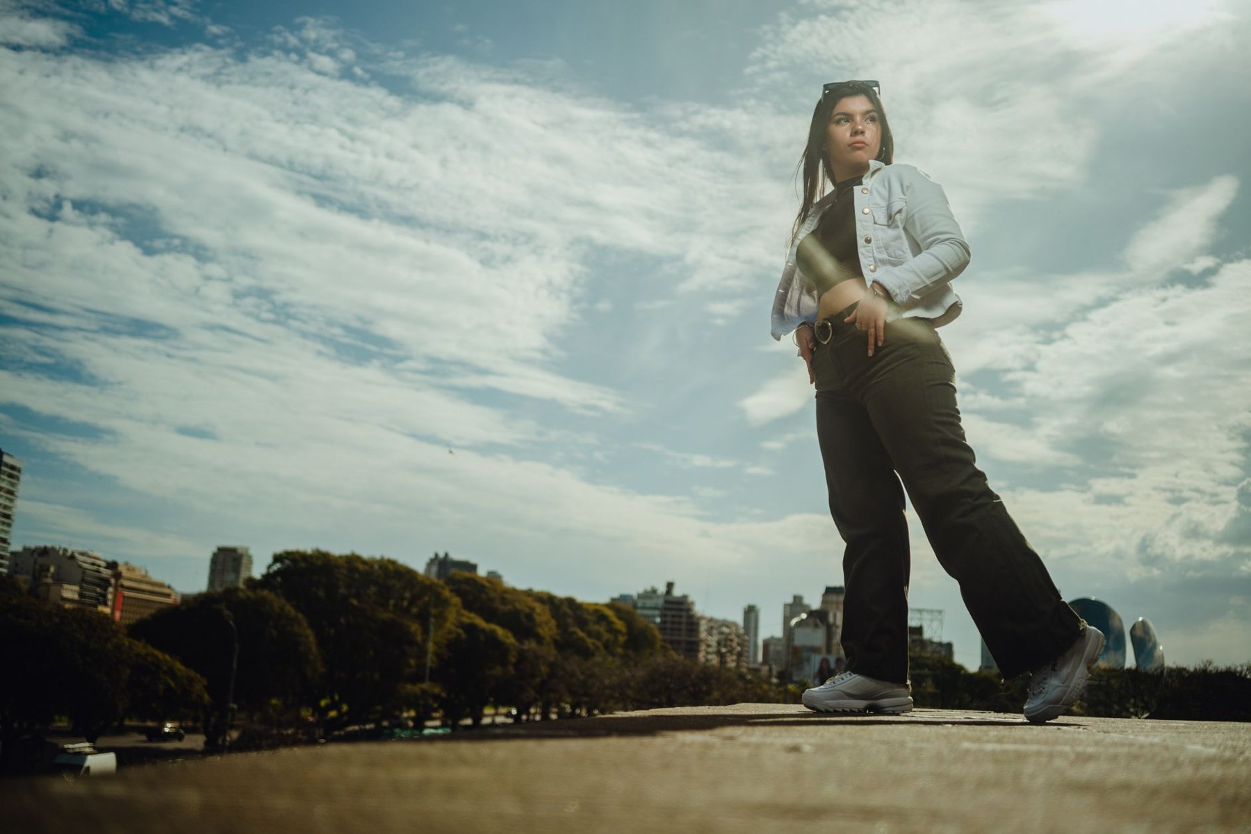 A woman wearing a light denim jacket, loose dark trousers, and sneakers stands outdoors beneath a cloudy sky.