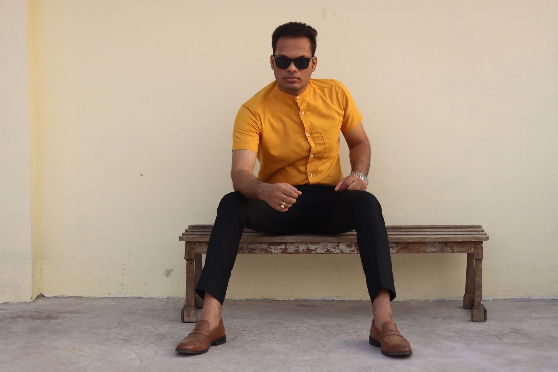 A man wearing a yellow short-sleeved shirt, cropped black trousers, brown loafers, and sunglasses sits on a wooden bench.