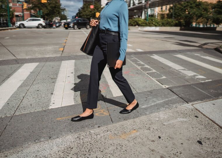 A person in a blue long-sleeved top, dark ankle-length trousers and black flats carries a shoulder bag across a city crosswalk.
