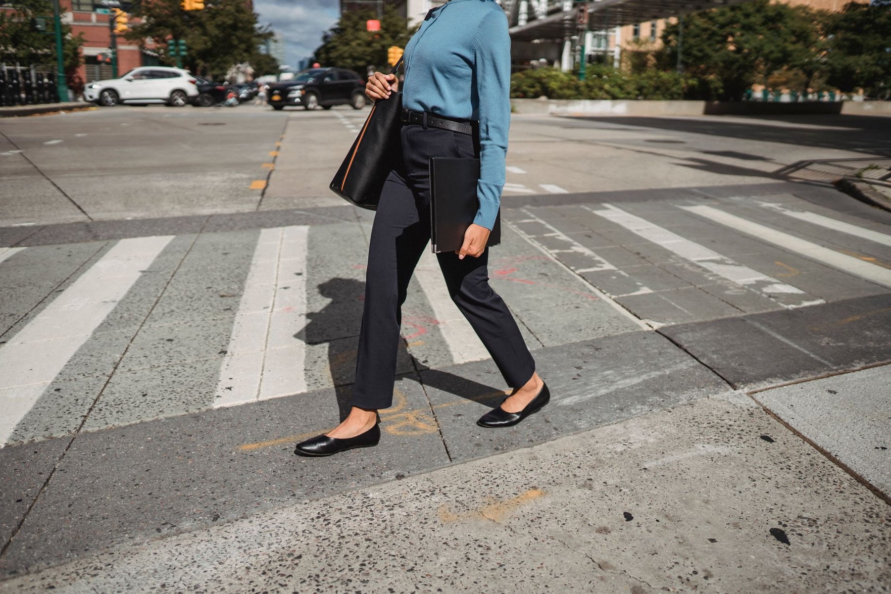 A person in a blue long-sleeved top, dark ankle-length trousers and black flats carries a shoulder bag across a city crosswalk.