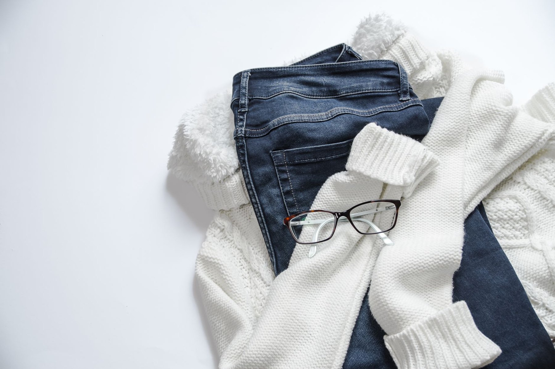 Dark blue jeans, white knitwear, and eyeglasses arranged on a light background.