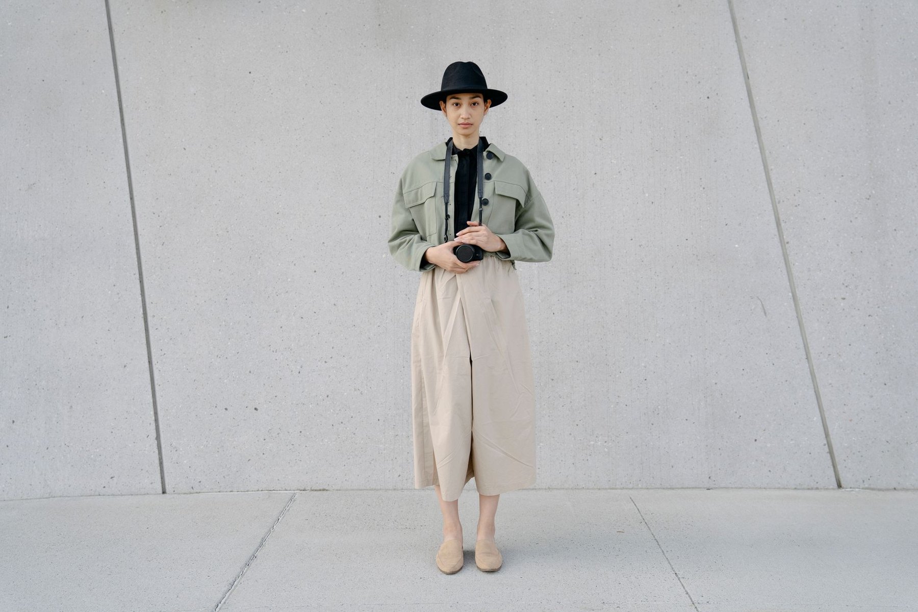 Woman wearing a short sage jacket, beige cropped wide-leg trousers, flats, and a black hat against a concrete wall.