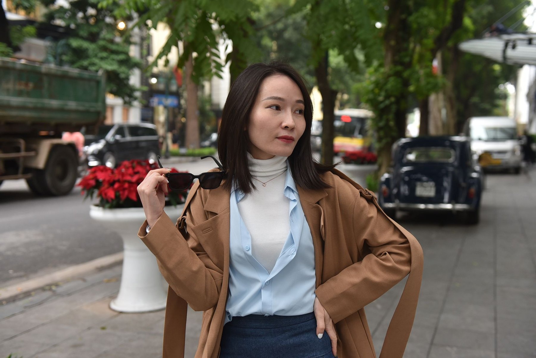 Woman on a city sidewalk wearing a brown coat over a pale blue shirt and white turtleneck, holding sunglasses.
