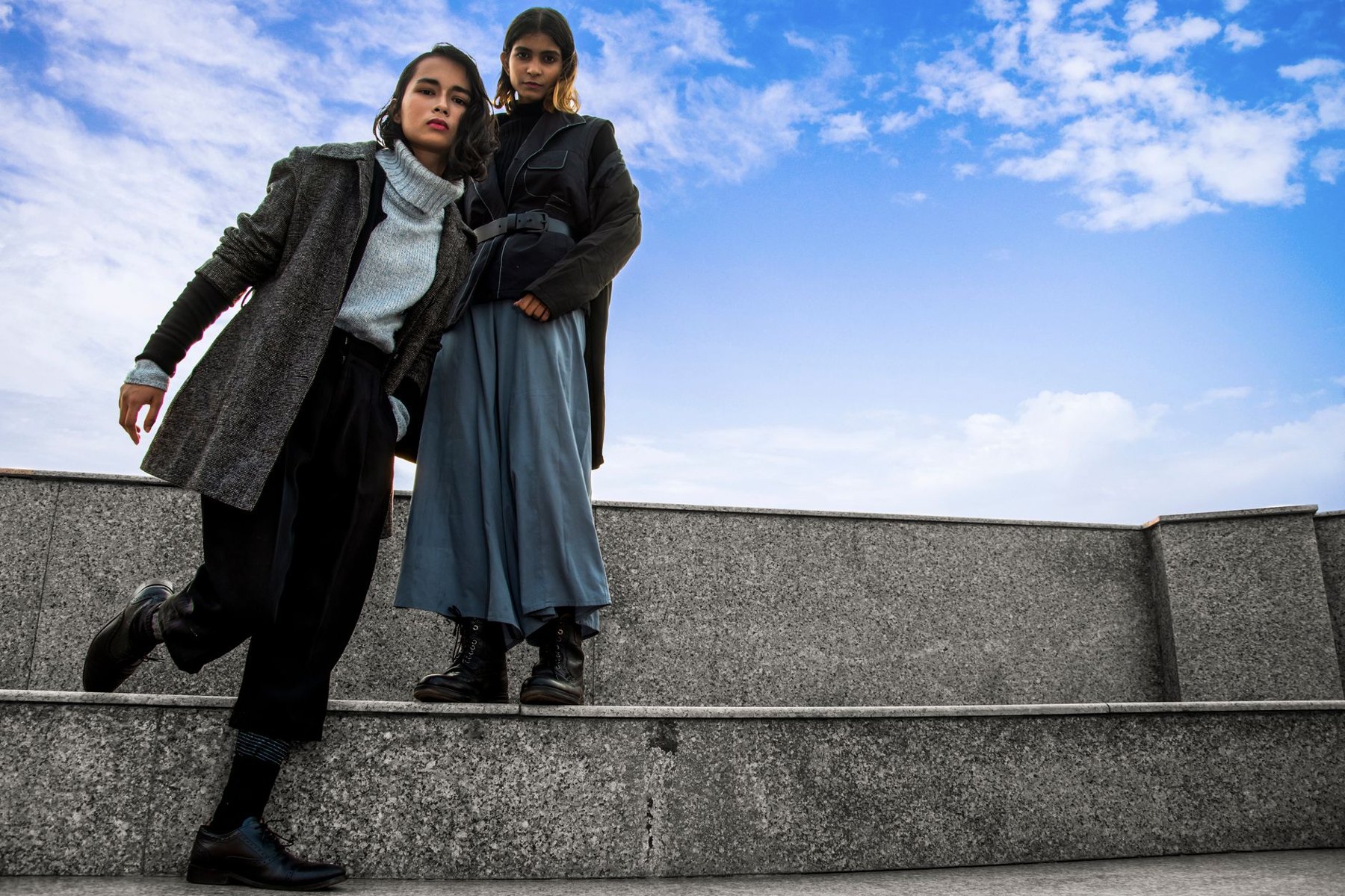 Two women pose on stone steps, one wearing a gray coat with black trousers and boots, the other a dark coat with a blue midi skirt and lace-up boots.