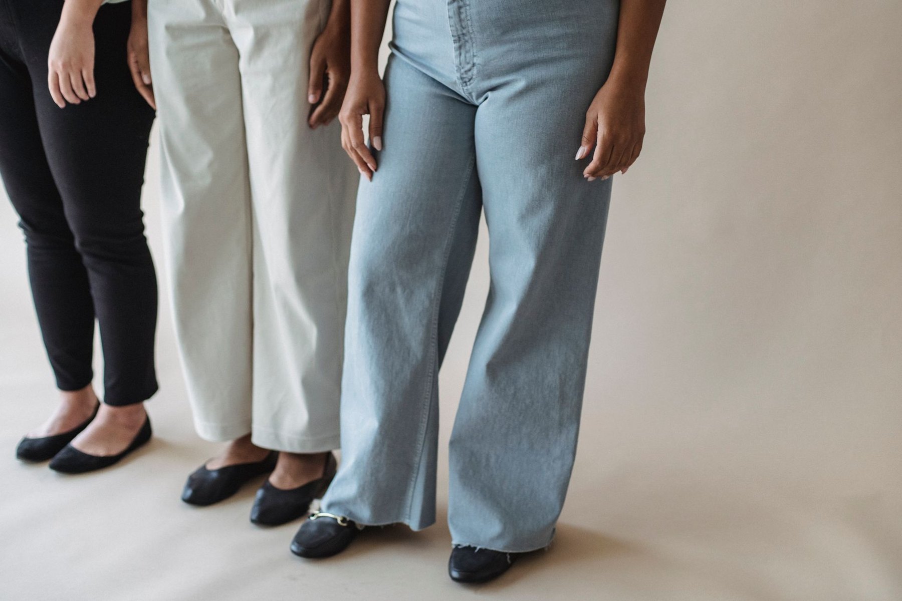 Three people wearing black trousers, cream trousers, and light blue jeans with dark flats against a neutral backdrop.