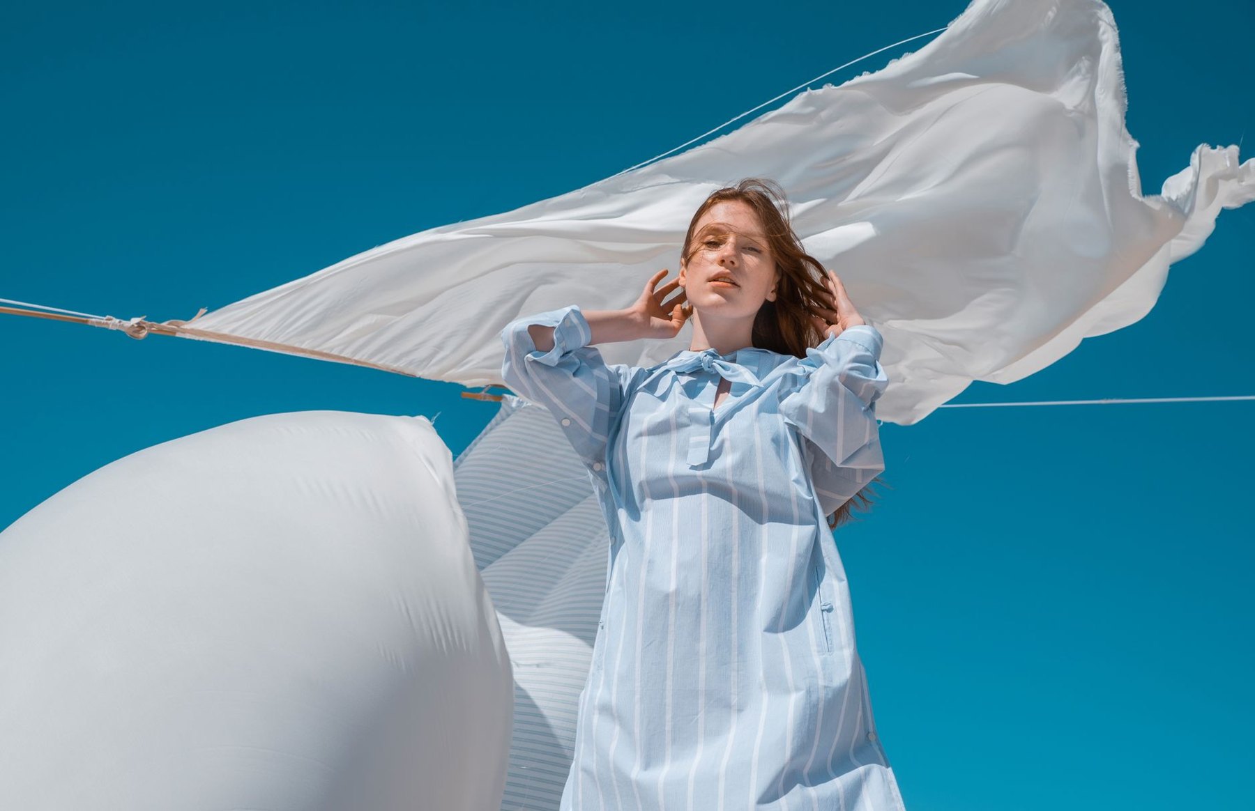A woman in a loose, light blue striped dress with long sleeves stands beneath billowing white sheets against a blue sky.