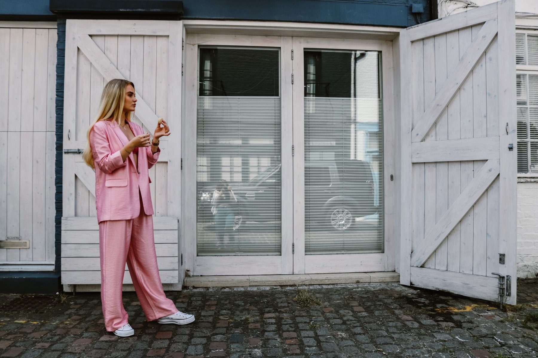 A woman in a loose pink blazer and wide-leg trousers wears white sneakers outside white wooden doors.