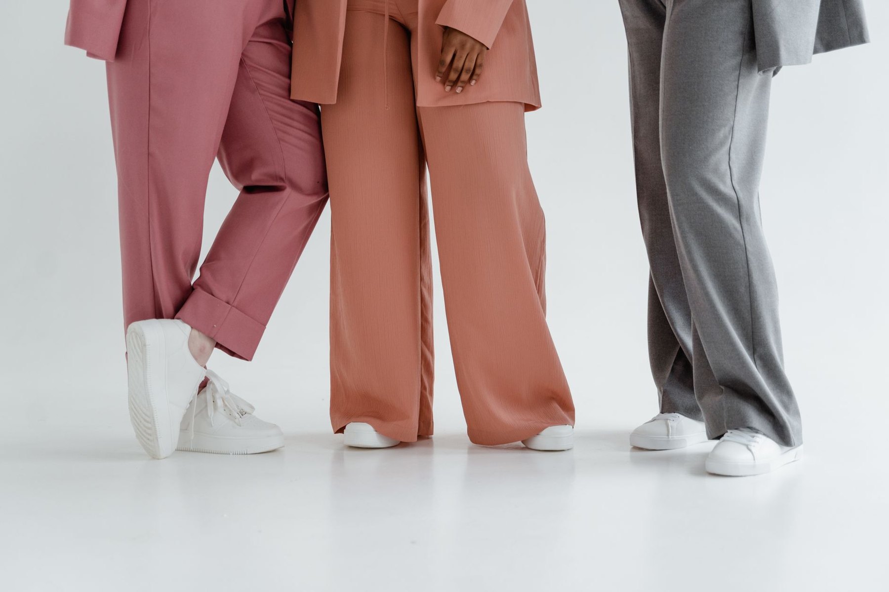 Three people wearing pink, peach, and gray trousers with white sneakers against a white studio background.