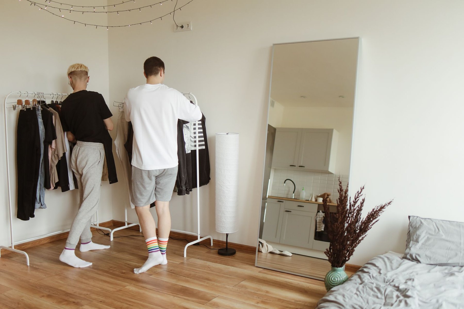 Two men look through clothes on a rack in a bedroom beside a full-length mirror.